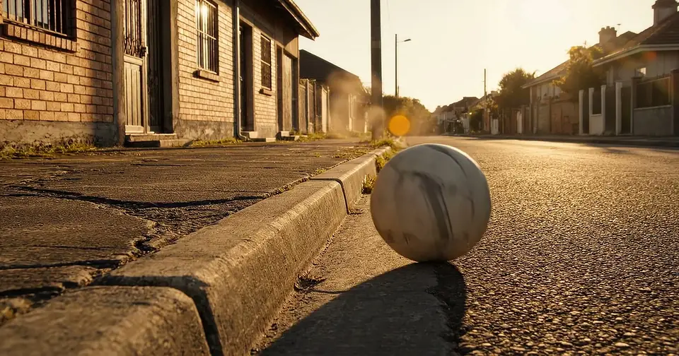 An old, worn football at rest against a kerb on an empty street at low sun.