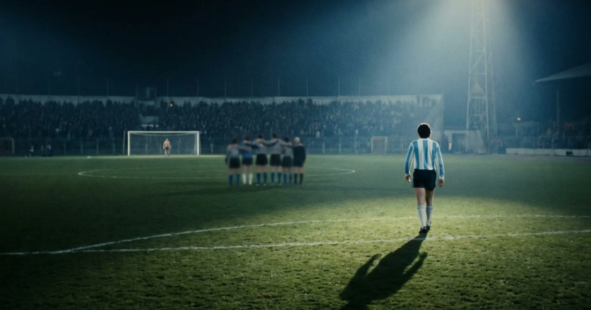 A lone player in pale blue and white stripes walking towards a distant goal and a huddled team, on a blue-lit pitch at night.