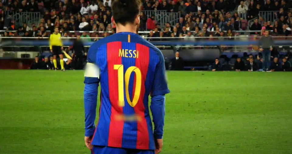 Messi seen from behind in a Barcelona shirt with a yellow 10, stands and a linesman ahead. Sponsor blurred in the film.