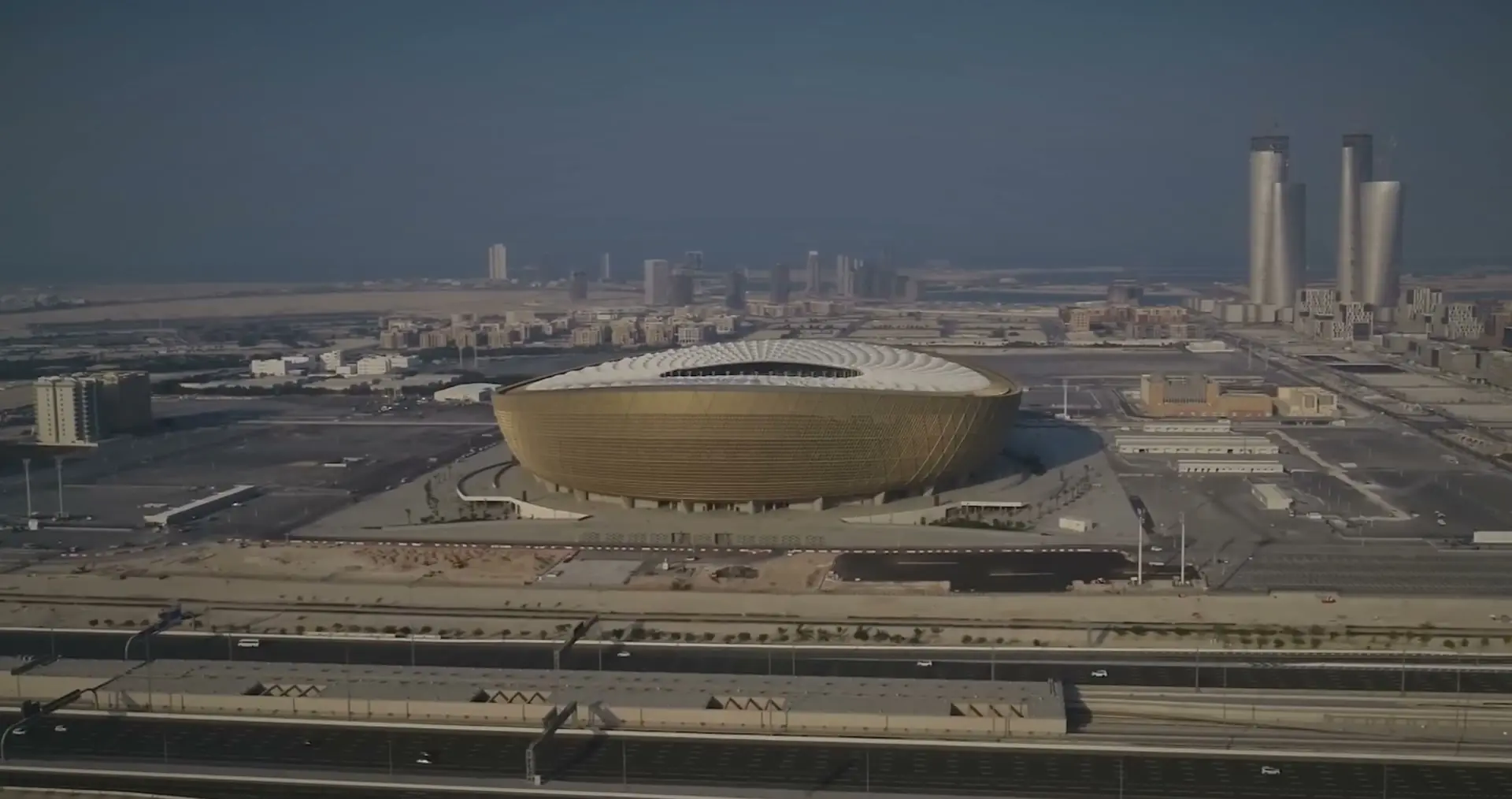 Aerial view of Lusail Stadium, Qatar, in daylight haze: a gold bowl in flat land, with towers on the horizon.