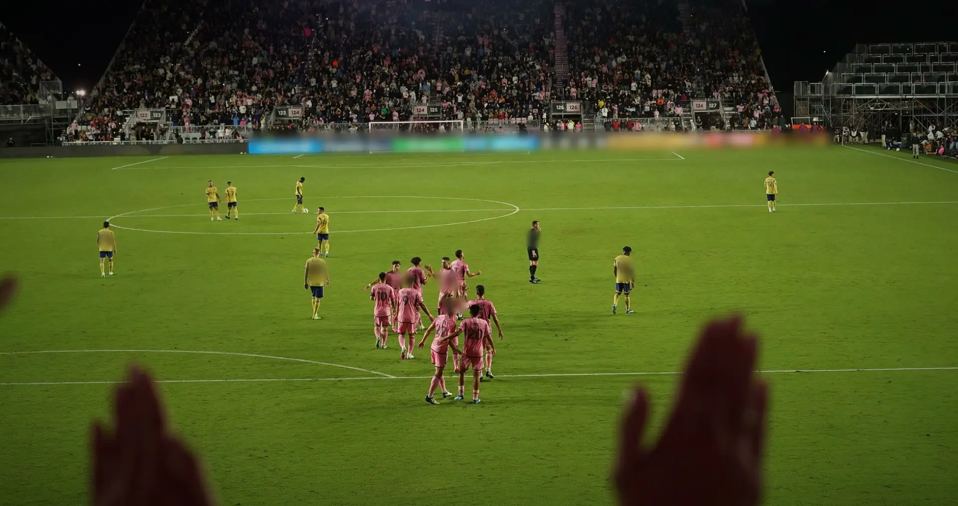 Inter Miami players in pink gathered on the pitch at night, visiting players in yellow around them, raised hands silhouetted in the foreground. Inter Miami v Real Salt Lake. Boards blurred in the film.