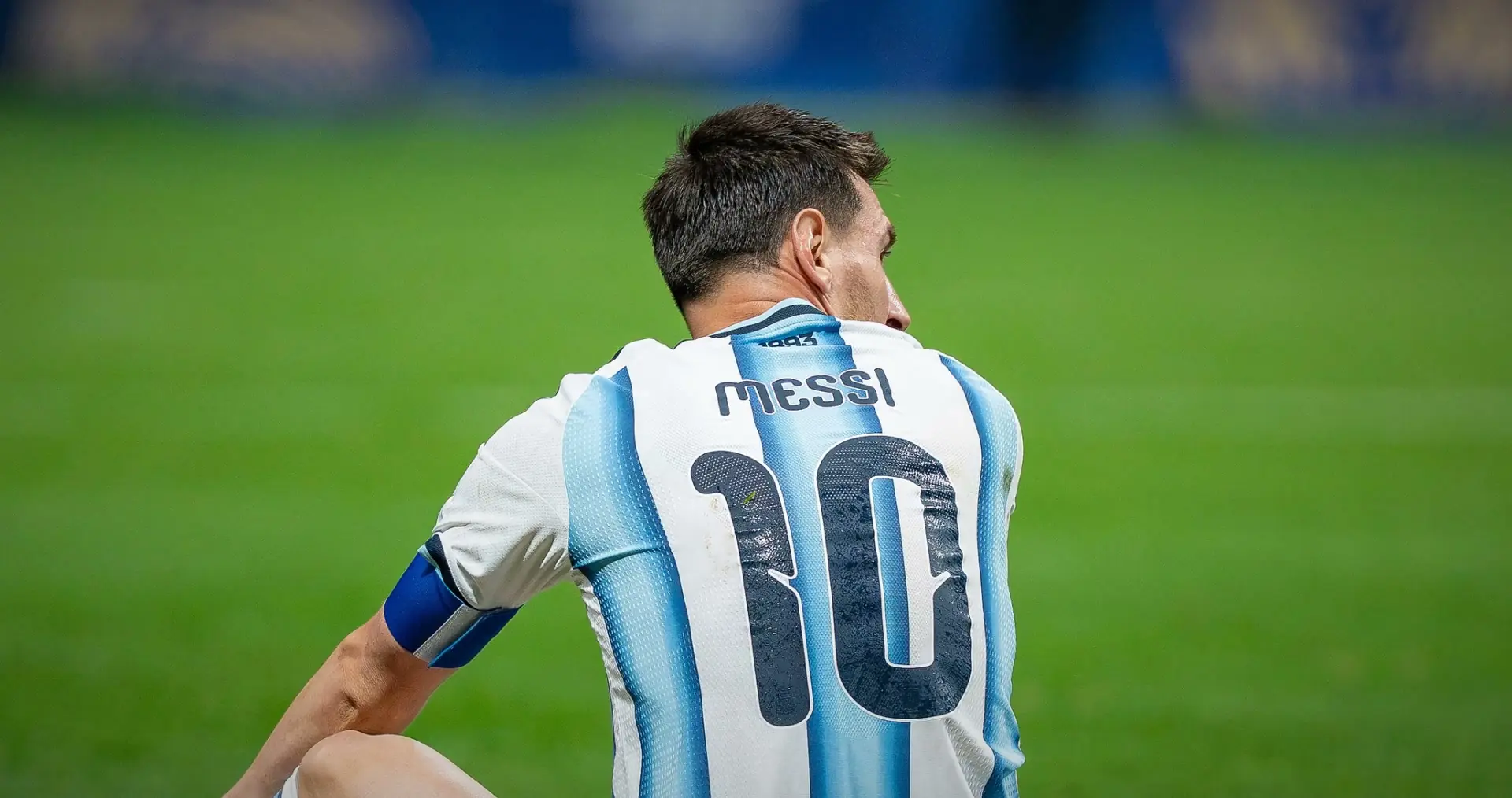 Messi seated on the grass, seen from behind and turning his head, in Argentina's 10 shirt, against plain green. Argentina v Egypt, 7 July 2026.