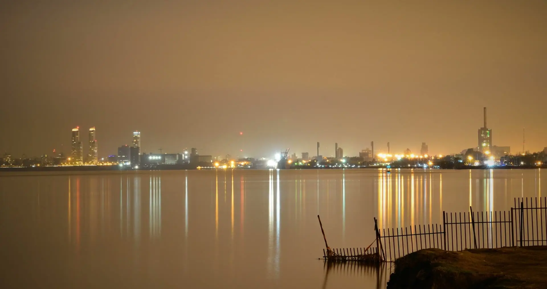The Rosario riverfront at night, with lights across the water under an orange sky and a broken fence in the foreground.