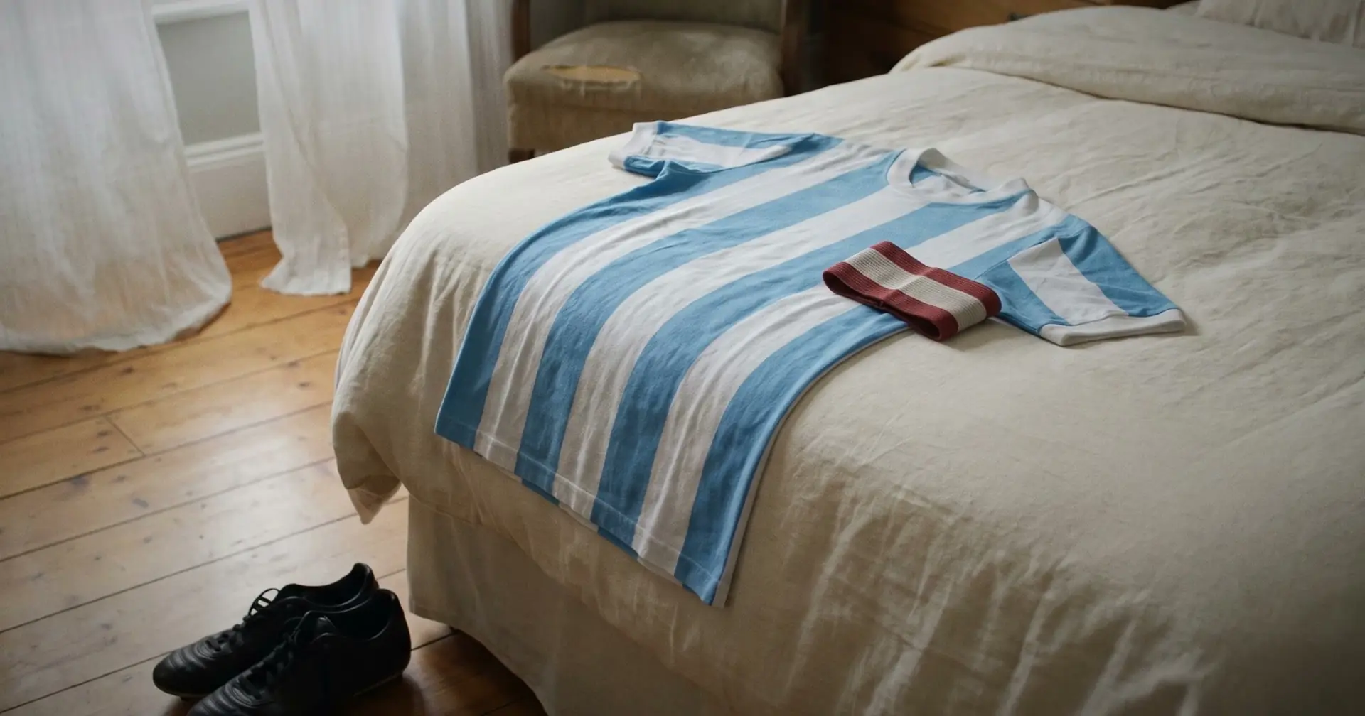 A striped pale blue and white shirt and a folded armband on a bed in a sunlit room, with boots on the floorboards.